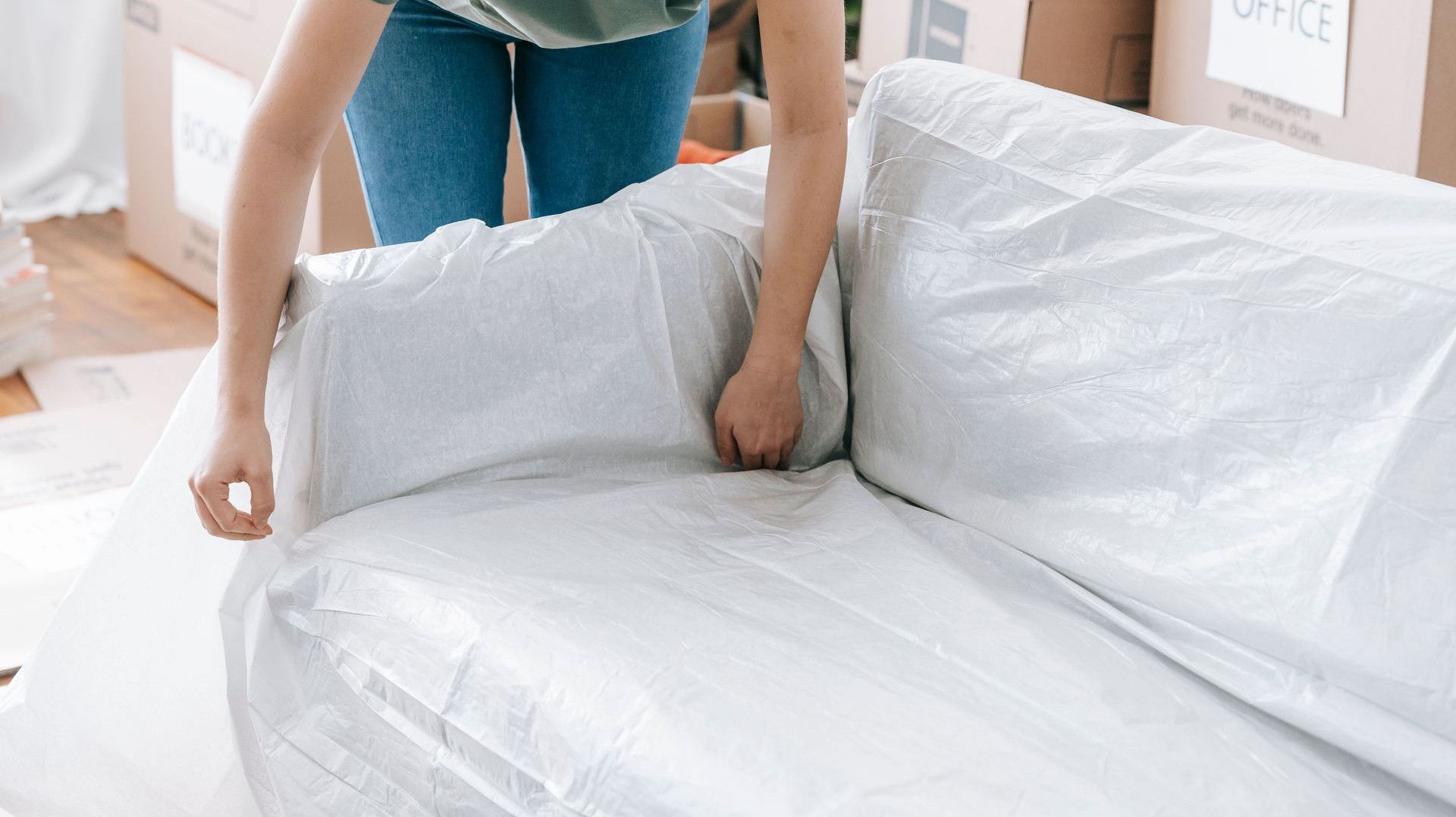 A woman carefully wraps a sofa in protective material while packing boxes in a bright room.